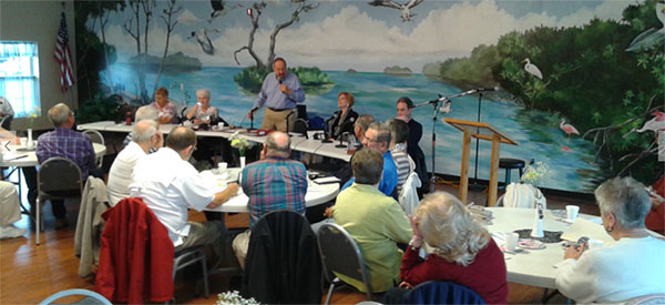 Senior man standing at a table, using a microphone to speak to a room of seniors during a meeting.