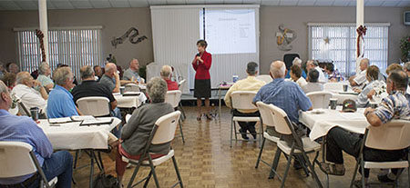 A woman in red jacket speaking a microphone to a room of seniors.