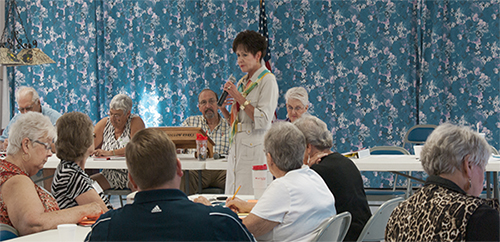 A woman speaking on a microphone while a group of seniors listen.